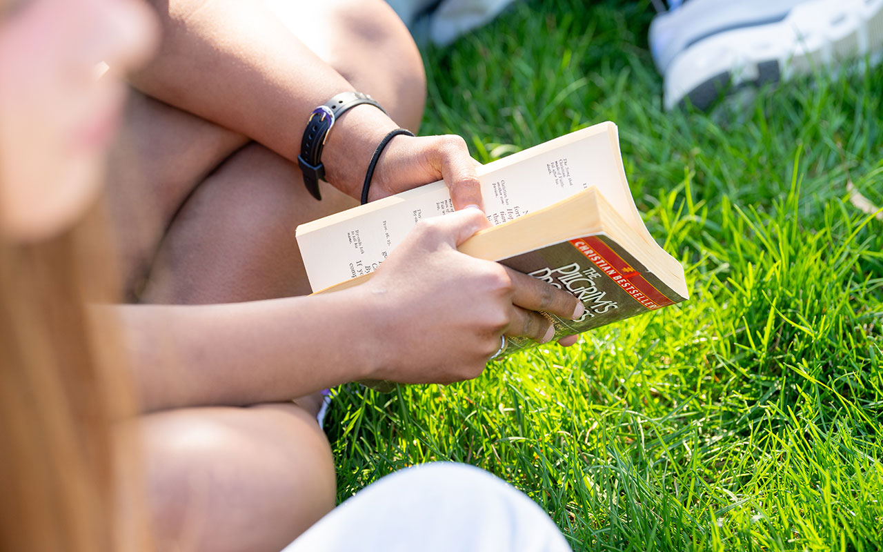 Student holding Pilgrim's Progress while sitting with other students on the grass.